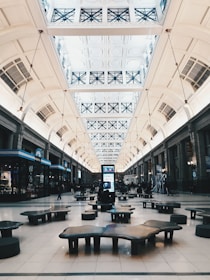 A spacious indoor hall features high ceilings with decorative panels and large skylights, allowing natural light to illuminate the area. The hall is lined with shops on either side, and modern seating areas are strategically placed throughout. A digital information board stands prominently in the center, while people are visible walking and seated around the space.
