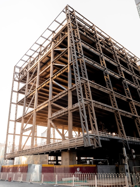 A partially constructed building with a visible steel framework, consisting of multiple floors and structural beams. The sunlight casts shadows on the metal surface, highlighting its industrial design. The foreground features a temporary fence and barriers, indicating an active construction site.