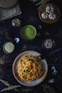 A wooden plate of spaghetti garnished with chopped herbs and sliced red chili peppers is placed on a dark surface. Surrounding the dish are various ingredients, including cloves of garlic, whole spices, a small bowl of creamy green soup, and a jar of seasoning. The setup is enhanced by rustic elements like a burlap cloth and dried flora, creating a cozy, earthy ambiance.