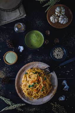 A wooden plate of spaghetti garnished with chopped herbs and sliced red chili peppers is placed on a dark surface. Surrounding the dish are various ingredients, including cloves of garlic, whole spices, a small bowl of creamy green soup, and a jar of seasoning. The setup is enhanced by rustic elements like a burlap cloth and dried flora, creating a cozy, earthy ambiance.
