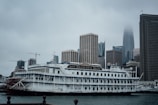A vintage-style riverboat, named San Francisco Belle, is docked in front of a modern city skyline under overcast skies. The boat features multiple decks, intricate railings, and a paddle wheel at the rear. Tall skyscrapers rise in the background, partially obscured by mist and low clouds.