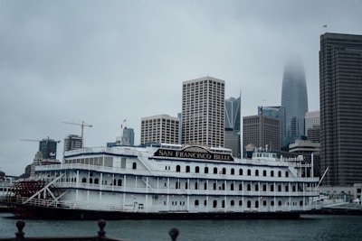 A vintage-style riverboat, named San Francisco Belle, is docked in front of a modern city skyline under overcast skies. The boat features multiple decks, intricate railings, and a paddle wheel at the rear. Tall skyscrapers rise in the background, partially obscured by mist and low clouds.