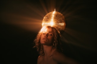 A person with curly hair is illuminated by the warm glow of a golden disco ball, casting an enchanting light pattern in the dark background. The lighting creates a dramatic and glamorous effect, highlighting their facial features and the texture of the disco ball.