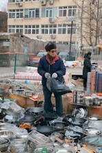 An outdoor market scene with a vendor standing among a variety of kitchen utensils. The vendor is wearing a blue jacket, gloves, and a hat, and is holding a black bag. There are stacks of pots, pans, and other metal cookware displayed around him on boxes and the ground. In the background, a beige building with numerous windows and air conditioning units is visible.