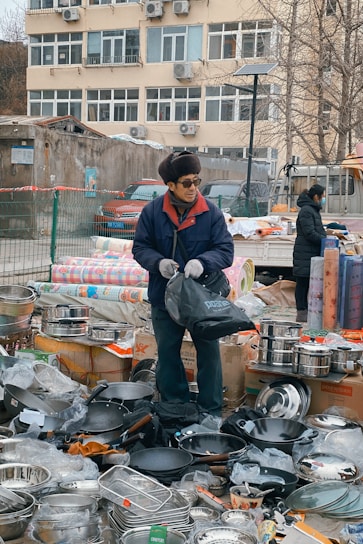 An outdoor market scene with a vendor standing among a variety of kitchen utensils. The vendor is wearing a blue jacket, gloves, and a hat, and is holding a black bag. There are stacks of pots, pans, and other metal cookware displayed around him on boxes and the ground. In the background, a beige building with numerous windows and air conditioning units is visible.