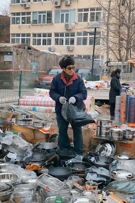An outdoor market scene with a vendor standing among a variety of kitchen utensils. The vendor is wearing a blue jacket, gloves, and a hat, and is holding a black bag. There are stacks of pots, pans, and other metal cookware displayed around him on boxes and the ground. In the background, a beige building with numerous windows and air conditioning units is visible.