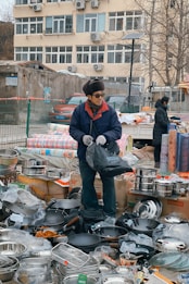 An outdoor market scene with a vendor standing among a variety of kitchen utensils. The vendor is wearing a blue jacket, gloves, and a hat, and is holding a black bag. There are stacks of pots, pans, and other metal cookware displayed around him on boxes and the ground. In the background, a beige building with numerous windows and air conditioning units is visible.