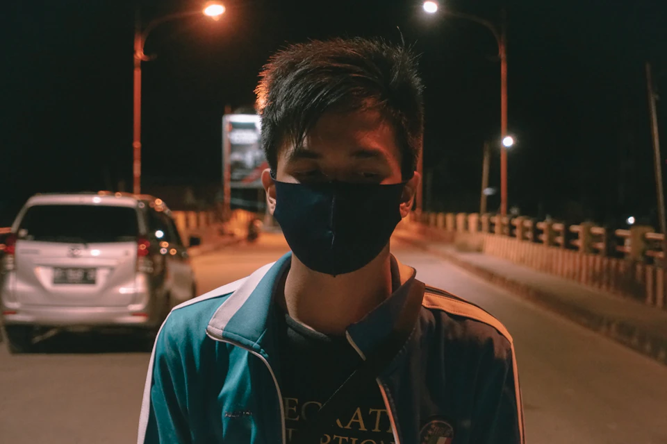 The image depicts a young man wearing a blue and white jacket, standing on a street at night with his head down. He is also wearing a face mask over his mouth and nose. In front of him, there are two cars parked - one white car to the left and another silver car further back in the scene. The background features a bridge illuminated by street lamps, creating an urban nighttime setting.