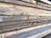 Multiple stacks of rough, unfinished wooden planks are piled horizontally, showcasing their natural grain and texture. The edges of the wood are uneven, with visible splinters and variations in color.