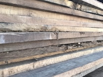 Multiple stacks of rough, unfinished wooden planks are piled horizontally, showcasing their natural grain and texture. The edges of the wood are uneven, with visible splinters and variations in color.