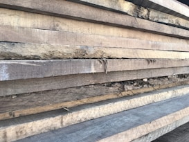 Multiple stacks of rough, unfinished wooden planks are piled horizontally, showcasing their natural grain and texture. The edges of the wood are uneven, with visible splinters and variations in color.