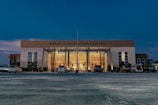 A large, modern building with the name 'AIMAS CONVENTION CENTRE' displayed prominently at the top. The structure features a glass entrance surrounded by tall columns, and several cars are parked in front on a spacious concrete area. The evening sky casts a deep blue hue over the scene, with soft lighting illuminating the entrance.