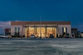 A large, modern building with the name 'AIMAS CONVENTION CENTRE' displayed prominently at the top. The structure features a glass entrance surrounded by tall columns, and several cars are parked in front on a spacious concrete area. The evening sky casts a deep blue hue over the scene, with soft lighting illuminating the entrance.