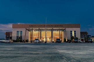 A large, modern building with the name 'AIMAS CONVENTION CENTRE' displayed prominently at the top. The structure features a glass entrance surrounded by tall columns, and several cars are parked in front on a spacious concrete area. The evening sky casts a deep blue hue over the scene, with soft lighting illuminating the entrance.
