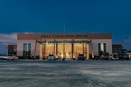 A large, modern building with the name 'AIMAS CONVENTION CENTRE' displayed prominently at the top. The structure features a glass entrance surrounded by tall columns, and several cars are parked in front on a spacious concrete area. The evening sky casts a deep blue hue over the scene, with soft lighting illuminating the entrance.