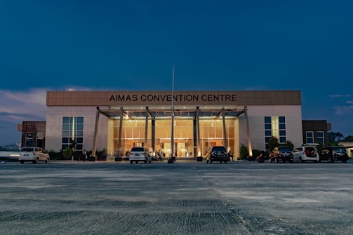 A large, modern building with the name 'AIMAS CONVENTION CENTRE' displayed prominently at the top. The structure features a glass entrance surrounded by tall columns, and several cars are parked in front on a spacious concrete area. The evening sky casts a deep blue hue over the scene, with soft lighting illuminating the entrance.