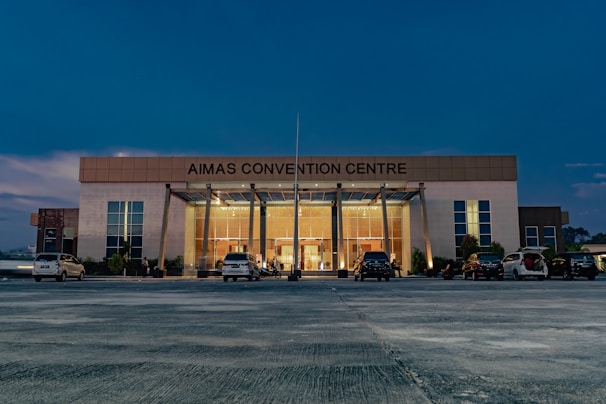 A large, modern building with the name 'AIMAS CONVENTION CENTRE' displayed prominently at the top. The structure features a glass entrance surrounded by tall columns, and several cars are parked in front on a spacious concrete area. The evening sky casts a deep blue hue over the scene, with soft lighting illuminating the entrance.