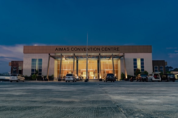 A large, modern building with the name 'AIMAS CONVENTION CENTRE' displayed prominently at the top. The structure features a glass entrance surrounded by tall columns, and several cars are parked in front on a spacious concrete area. The evening sky casts a deep blue hue over the scene, with soft lighting illuminating the entrance.