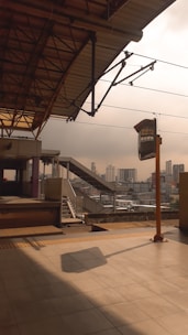 A train station platform with an overhead roof structure is depicted. The platform features stairs and a railing leading to an upper area. In the background, a city skyline with multiple buildings is visible under an overcast sky. A signpost stands near the platform edge, casting a shadow on the tiled floor.