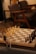 A chessboard set up for a game with white and dark chess pieces positioned for play. The setting includes soft lighting, a wooden table, and comfortable chairs in the background, creating a cozy atmosphere. A small vase with dried flowers is seen to the side of the chessboard.