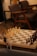A chessboard set up for a game with white and dark chess pieces positioned for play. The setting includes soft lighting, a wooden table, and comfortable chairs in the background, creating a cozy atmosphere. A small vase with dried flowers is seen to the side of the chessboard.