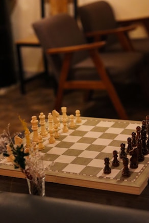 A chessboard set up for a game with white and dark chess pieces positioned for play. The setting includes soft lighting, a wooden table, and comfortable chairs in the background, creating a cozy atmosphere. A small vase with dried flowers is seen to the side of the chessboard.