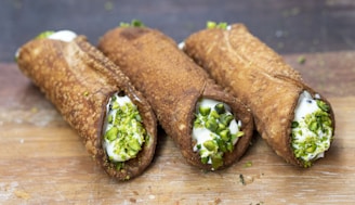 Three traditional cannoli are arranged on a wooden surface. The crispy, golden-brown pastry shells are filled with a creamy ricotta mixture, and each end is garnished with chopped pistachios.