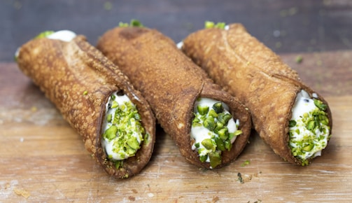Three traditional cannoli are arranged on a wooden surface. The crispy, golden-brown pastry shells are filled with a creamy ricotta mixture, and each end is garnished with chopped pistachios.