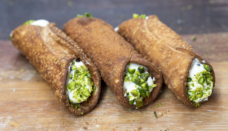 Three traditional cannoli are arranged on a wooden surface. The crispy, golden-brown pastry shells are filled with a creamy ricotta mixture, and each end is garnished with chopped pistachios.