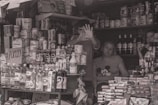 A small cluttered shop with shelves full of various products such as cleaning supplies, baby formula, and bottled beverages. In the center, a person is making a hand gesture towards the camera. Two other people are behind the counter, one looking at the camera and the other seemingly engaged in a task.