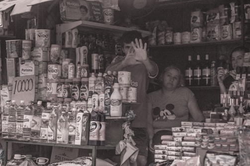 A small cluttered shop with shelves full of various products such as cleaning supplies, baby formula, and bottled beverages. In the center, a person is making a hand gesture towards the camera. Two other people are behind the counter, one looking at the camera and the other seemingly engaged in a task.