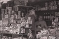 A small cluttered shop with shelves full of various products such as cleaning supplies, baby formula, and bottled beverages. In the center, a person is making a hand gesture towards the camera. Two other people are behind the counter, one looking at the camera and the other seemingly engaged in a task.