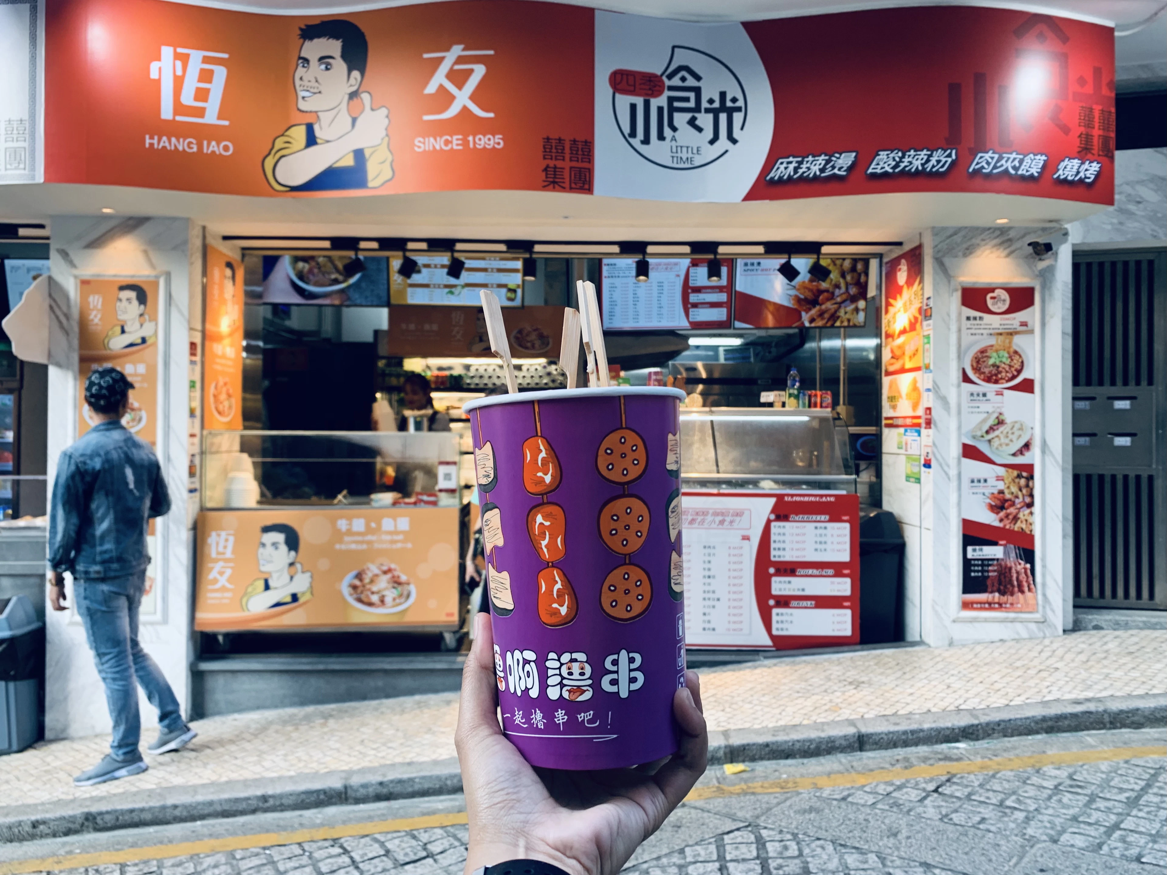 A cup of skewered street snacks held up in front of a snack shop on Avenida de Almeida Ribeiro in Macau
