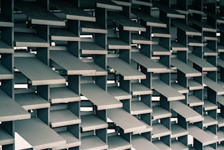 A geometric structure composed of stacked, rectangular concrete slabs arranged in a grid pattern. These slabs are layered at various angles, creating a dynamic and visually intriguing lattice design. The background hints at an urban environment visible through the gaps between the slabs.