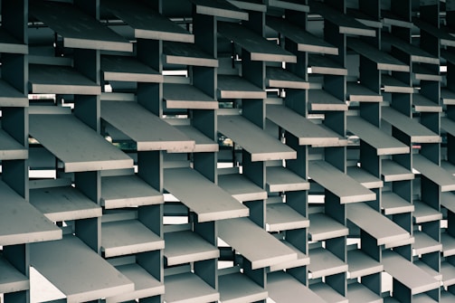 A geometric structure composed of stacked, rectangular concrete slabs arranged in a grid pattern. These slabs are layered at various angles, creating a dynamic and visually intriguing lattice design. The background hints at an urban environment visible through the gaps between the slabs.