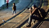 A photographer sits on stone steps near a body of water, focusing his camera on a pigeon walking nearby. Two children stand on the walkway, one looking towards the water and the other with hands in pockets. The scene is warmly lit by the sun, casting long shadows.