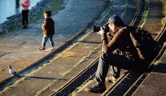 A photographer sits on stone steps near a body of water, focusing his camera on a pigeon walking nearby. Two children stand on the walkway, one looking towards the water and the other with hands in pockets. The scene is warmly lit by the sun, casting long shadows.