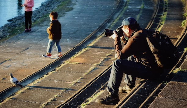 A photographer sits on stone steps near a body of water, focusing his camera on a pigeon walking nearby. Two children stand on the walkway, one looking towards the water and the other with hands in pockets. The scene is warmly lit by the sun, casting long shadows.