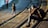 A photographer sits on stone steps near a body of water, focusing his camera on a pigeon walking nearby. Two children stand on the walkway, one looking towards the water and the other with hands in pockets. The scene is warmly lit by the sun, casting long shadows.