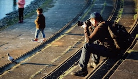 A photographer sits on stone steps near a body of water, focusing his camera on a pigeon walking nearby. Two children stand on the walkway, one looking towards the water and the other with hands in pockets. The scene is warmly lit by the sun, casting long shadows.