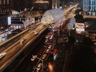 A bustling urban scene at night featuring a multi-lane elevated highway with fast-moving vehicles. Below the highway are city streets with cars and pedestrians. Buildings with illuminated signs are visible, contributing to a vibrant city atmosphere. A glass-covered structure runs parallel to the highway, likely a public transportation hub.