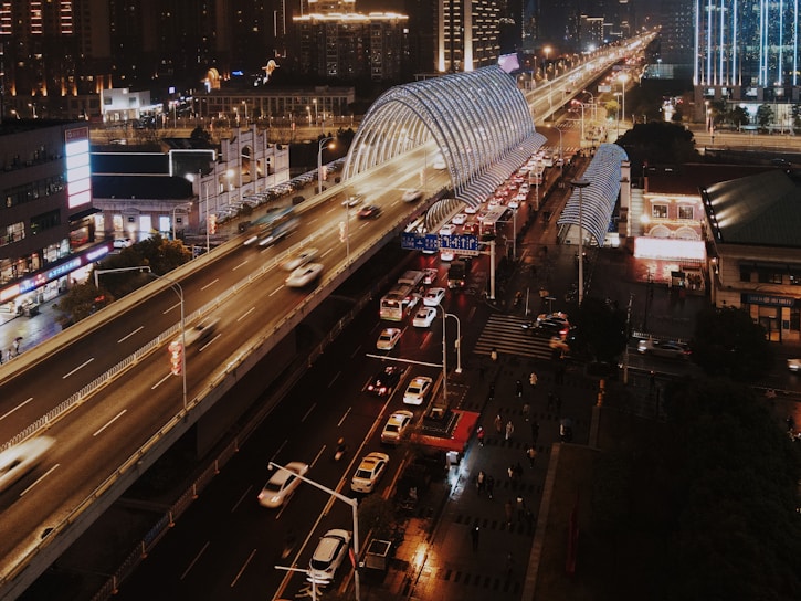 A bustling urban scene at night featuring a multi-lane elevated highway with fast-moving vehicles. Below the highway are city streets with cars and pedestrians. Buildings with illuminated signs are visible, contributing to a vibrant city atmosphere. A glass-covered structure runs parallel to the highway, likely a public transportation hub.