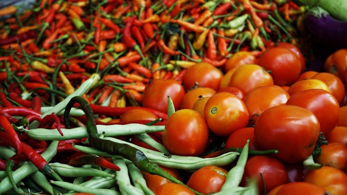 A vibrant assortment of fresh vegetables including red tomatoes, green beans, and a variety of colorful chili peppers. The produce is piled high, showcasing a variety of shapes and textures.