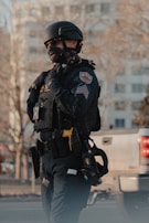A police officer in tactical gear, including a helmet and protective vest, stands in an urban environment. The officer is holding a radio microphone near their mouth, suggesting communication. The uniform features patches indicating their department and rank. In the background, a blurred landscape shows manufactured structures, but they are nondescript.