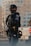 A police officer in tactical gear, including a helmet and protective vest, stands in an urban environment. The officer is holding a radio microphone near their mouth, suggesting communication. The uniform features patches indicating their department and rank. In the background, a blurred landscape shows manufactured structures, but they are nondescript.