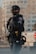 A police officer in tactical gear, including a helmet and protective vest, stands in an urban environment. The officer is holding a radio microphone near their mouth, suggesting communication. The uniform features patches indicating their department and rank. In the background, a blurred landscape shows manufactured structures, but they are nondescript.