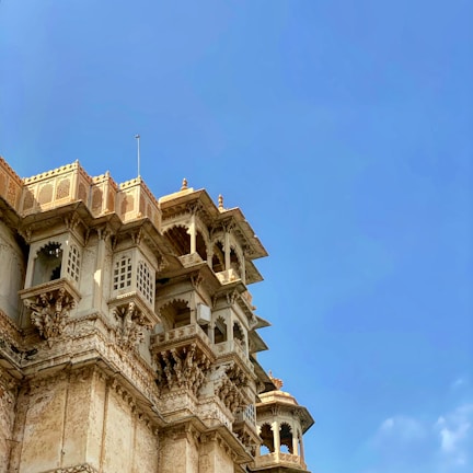 A detailed architectural structure with intricate carvings and designs adorns a corner of the building. The structure features ornate jharokhas and latticed windows, characteristic of traditional Indian architecture. The sky in the background is a vibrant blue, enhancing the aged elegance of the building.