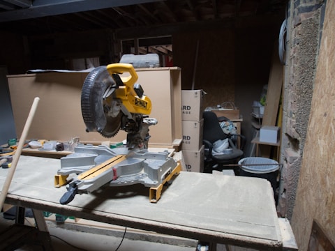 A cluttered workshop space features a miter saw positioned on a workbench. The saw is surrounded by various tools and materials, including cardboard boxes, an office chair, and a bin. The area is dimly lit, with exposed brick walls and wooden structures, suggesting ongoing construction or renovation work.