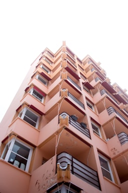 A tall, peach-colored apartment building viewed from a low angle. The building features multiple balconies with maroon railings and some visible air conditioning units. Parts of the building's facade show signs of wear and peeling paint. The sky above is overcast, contributing to a neutral lighting condition.