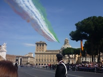 Jets fly overhead releasing smoke trails in the colors of the Italian flag against a clear blue sky, with an ornate historic building and a dome in the background. Trees and a crowd of people are visible, along with uniformed personnel in the foreground.